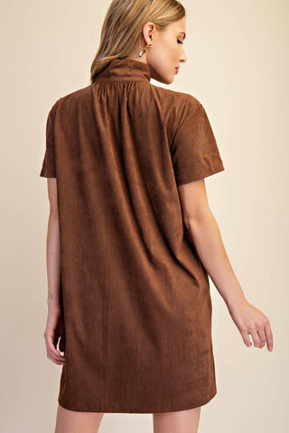A woman shown from the back wearing a brown short-sleeve dress with a gathered high neck, photographed against a plain beige studio background.