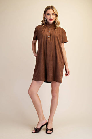 A woman modeling a brown short-sleeve mini dress with a high gathered neckline and decorative buttons, paired with dark brown heeled sandals, against a solid beige studio background.