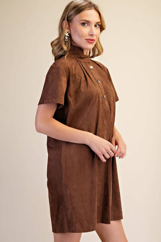 A woman modeling a brown short-sleeve mock-neck dress with a gathered front and decorative buttons, photographed against a plain beige studio background.