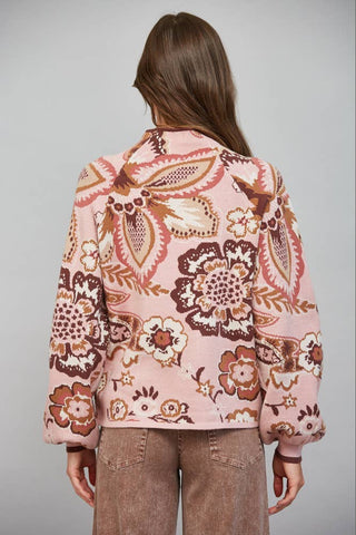 A woman viewed from behind wearing a light pink cropped jacket with a brown and beige floral paisley pattern, paired with brown pants, against a solid gray studio background.