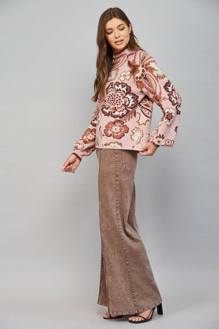 A woman modeling a pink long-sleeve knit top with a brown and beige floral paisley pattern, paired with brown wide-leg jeans and black heeled sandals, against a solid gray studio background.