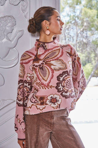 A woman wearing a pink long-sleeve mock-neck top with a brown and beige butterfly pattern, paired with brown corduroy pants, standing next to a white classical column.