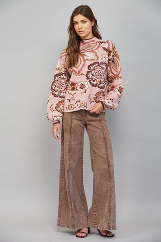 A woman modeling a pink long-sleeve top with a brown and beige paisley pattern, paired with brown wide-leg pants, against a solid gray studio background.