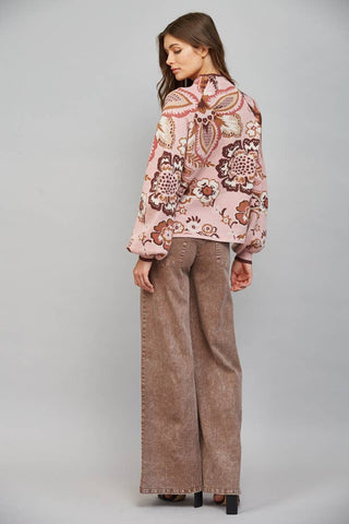 A woman stands in profile wearing a light pink cropped jacket with a brown and beige paisley pattern, paired with wide-leg brown pants, against a neutral gray studio background.