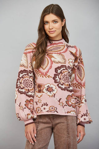 A woman modeling a pink long-sleeve knit top with a brown and beige floral paisley pattern, paired with brown pants, against a solid gray studio background.