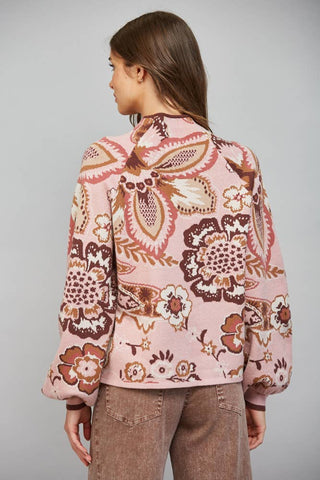A woman shown from the back wearing a pink long-sleeve bomber jacket with a brown and beige paisley and floral pattern, paired with brown pants, against a solid gray studio background.
