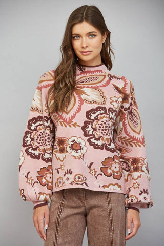 A woman modeling a pink long-sleeve mock neck sweater with a brown and beige floral paisley pattern, paired with brown pants, against a solid gray studio background.