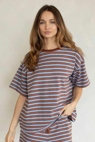A woman wearing an oversized short-sleeve t-shirt with horizontal brown and light blue stripes, photographed against a plain light beige wall.