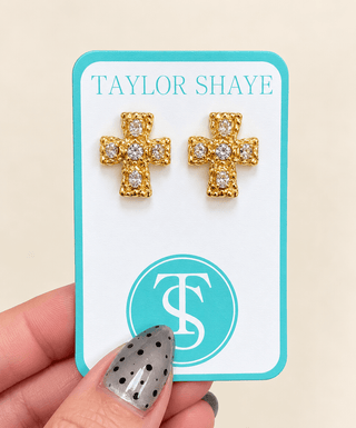 A pair of gold cross stud earrings with clear crystals, displayed on a white and turquoise card branded 'Taylor Shaye', held by a hand with a black-and-white polka dot nail.
