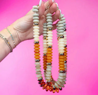 A hand holds up three beaded strands of mixed jewelry against a bright pink background. The beads include pale gray-green, cream, orange, amber, and metallic gold and silver accents.