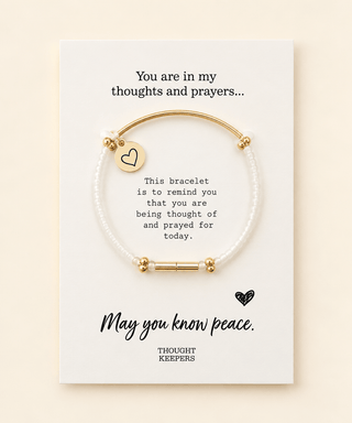 A white beaded bracelet with gold-toned metal accents and a small heart charm, displayed on a white printed card with sentimental text. The card reads, 'You are in my thoughts and prayers.