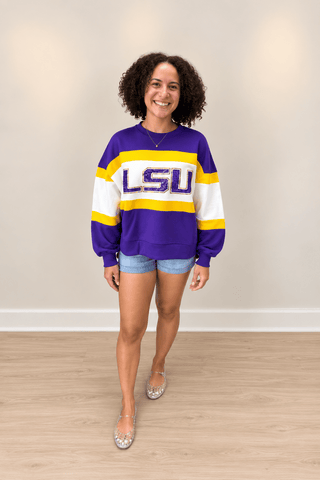 A woman modeling a purple, white, and yellow LSU crewneck sweatshirt, paired with denim shorts and silver flat shoes, standing in a room with a beige wall and light wood floor.