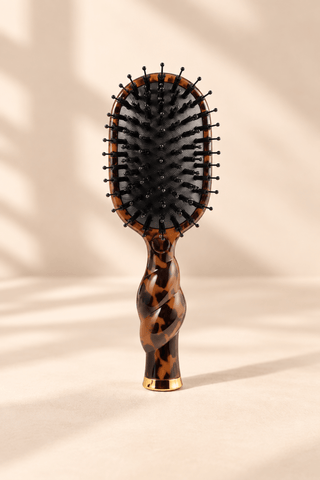 A classic oval hairbrush with a glossy tortoiseshell handle and black bristles, standing upright on a beige surface with soft leaf shadows.