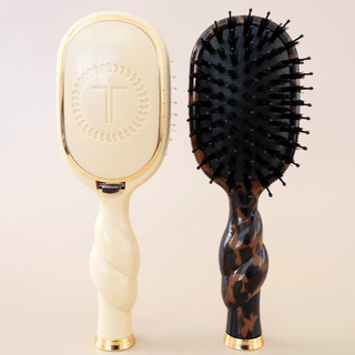 Two paddle hairbrushes shown side by side on a light beige studio background. The left brush has an oval cream face with gold trim and a twisted handle with gold accents.