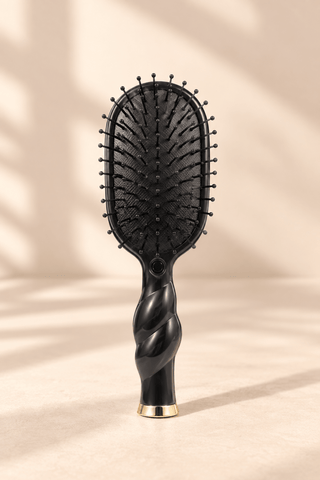 A black paddle hairbrush with a sculpted twisted handle and a gold accent at the base, standing upright on a beige surface with soft shadow overlays.