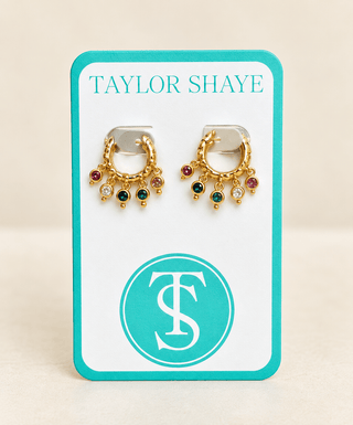 A pair of gold huggie hoop earrings with dangling colorful gemstone accents, displayed on a white and teal Taylor Shaye brand card against a light beige background.