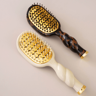 Two hairbrushes on a light beige background. One has a tortoiseshell pattern handle with black and brown tones, and the other has a cream-colored handle with gold accents.