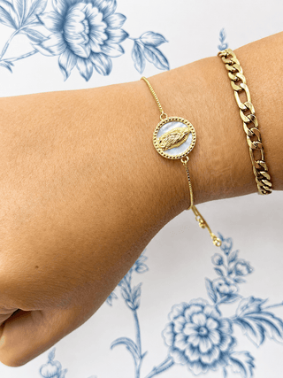 A close-up of a person's wrist wearing two gold bracelets: a delicate chain bracelet with a round white inlay and gold bird motif, and a thicker gold curb chain, against a white background with blue floral print.