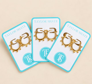 Three gold hoop earrings with small bead details, displayed on white cards with turquoise borders. The cards are branded 'Taylor Shaye' and arranged in a fan shape on a light beige background.