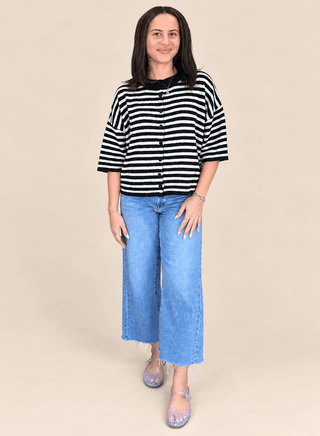 A woman modeling a black and white horizontally striped short-sleeve cardigan with a button-up front, paired with wide-leg light blue jeans and purple glittery flats, against a solid beige background.