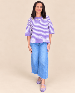 A woman modeling a lavender and white striped short-sleeve button-up top with blue wide-leg jeans and matching purple sandals, set against a solid beige studio background.