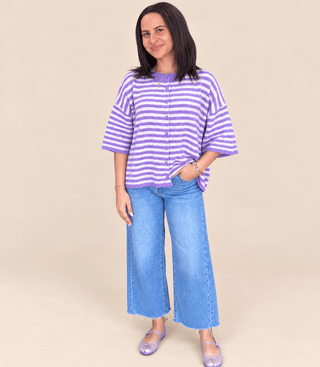 A woman modeling a purple and white horizontally striped short-sleeve button-up top with light blue wide-leg jeans and lavender flat shoes, standing against a solid beige studio background.