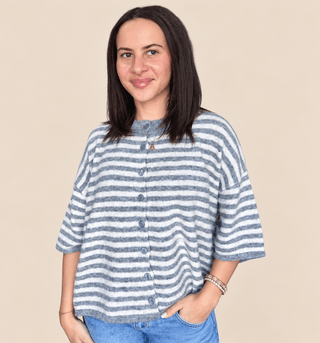 A woman with dark shoulder-length hair wearing a blue and white striped short-sleeve button-up top and blue jeans, standing against a solid beige background.