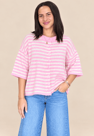 A woman modeling a pink and white striped short-sleeve button-up top with blue jeans, standing against a solid beige background.