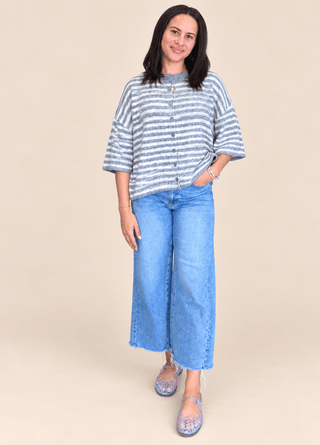 A woman modeling a light blue and white striped short-sleeve button-up shirt with wide-leg blue jeans and purple glittery flats, set against a solid beige studio background.
