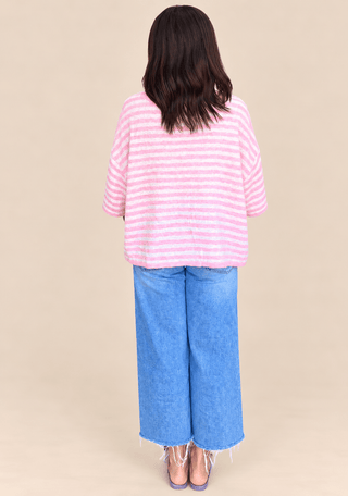 A woman shown from the back wearing a pink and white striped knit top, wide-leg blue jeans, and purple heels against a solid beige studio background.