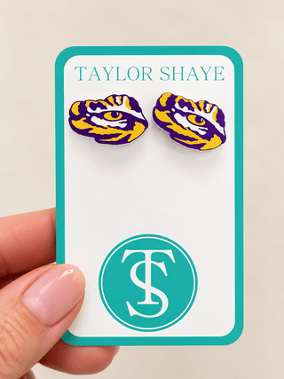 A hand holds a white display card with turquoise border, featuring a pair of tiger-eye stud earrings above the text 'TAYLOR SHAYE' and a circular TS logo. The earrings are purple, yellow, and white with a gold eye detail, representing the LSU Tigers.