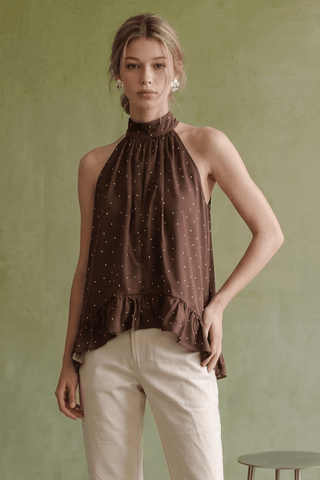 A woman modeling a brown halter-neck top with a small white polka dot pattern and a ruffled hem, paired with cream pants, set against a textured olive green wall.