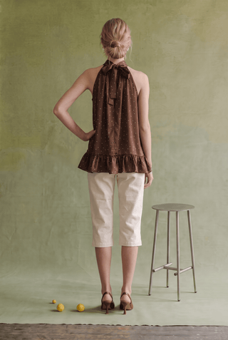 A woman stands with her back to the camera wearing a brown halter top with a ruffled hem, cream cropped pants, and brown heels, posed in front of a textured sage green backdrop with a metal stool and small yellow lemons on the floor.