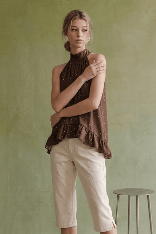A woman modeling a sleeveless high-neck brown top with small white polka dots and a ruffled hem, paired with light beige cropped pants, standing against a textured sage green wall.