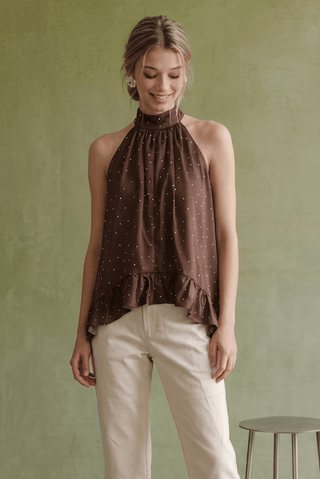A woman wearing a brown halter-neck top with a small white polka dot pattern and cream pants, standing against a textured sage green wall.