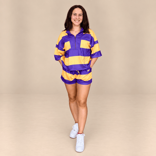 A woman modeling a matching purple and yellow striped athletic set consisting of a half-zip pullover and shorts, paired with white sneakers, against a solid beige studio background.