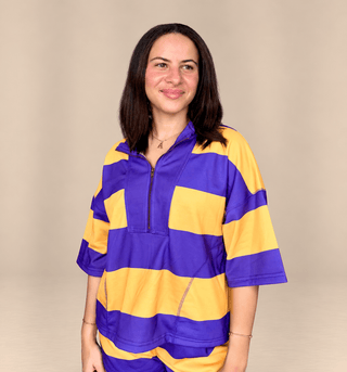 A woman wearing a purple and yellow striped athletic half-zip top, standing against a solid beige studio background.