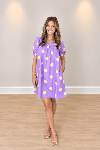 A woman modeling a lavender purple short-sleeve dress with yellow polka dots, standing in a room with a beige wall and light wood floor.