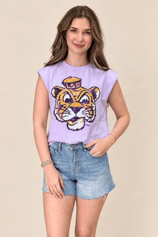 A woman modeling a lavender sleeveless crop top with a large embroidered tiger mascot graphic on the front, paired with light-wash denim shorts, against a solid beige background.