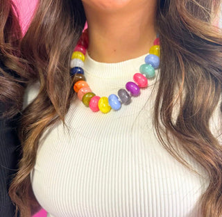A close-up of a person wearing a chunky multi-colored beaded necklace made of glossy, oval-shaped beads in various bright colors, paired with a cream ribbed top.