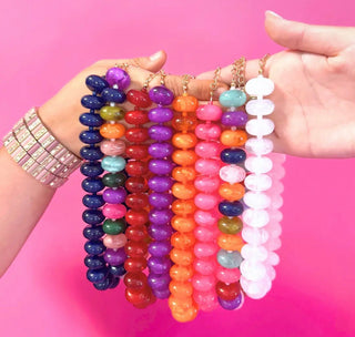 Six colorful beaded gemstone bracelets held up by two hands against a bright pink background. The bracelets feature round polished beads in various colors including purple, blue, red, orange, pink, and white, with gold chain links.