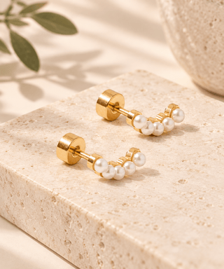 A pair of gold stud earrings with small white pearl clusters, displayed on a beige stone block with soft shadows and a blurred green leaf in the background.