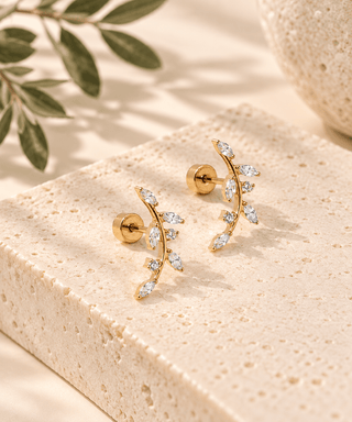 A pair of gold leaf-shaped stud earrings with clear marquise and oval gemstones, displayed on a textured beige stone block with soft-focus olive leaves in the background.