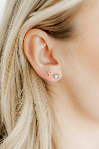 Close-up of an ear wearing gold stud earrings with a cross and heart design.