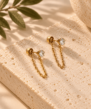 A pair of gold chain drop earrings with clear gemstones, displayed on a textured beige stone block with soft shadows and a leaf in the background.