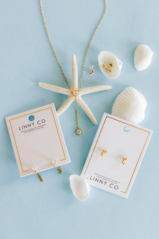 A flat-lay of jewelry from the brand LINNY CO on a light blue background, featuring a gold necklace with a starfish pendant and matching gold stud earrings on white display cards, styled with white seashells.