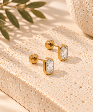 A pair of gold-toned rectangular stud earrings with clear marquise-cut stones, displayed on a textured beige stone block with soft shadows and green leaves in the background.