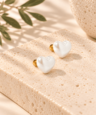 Two small heart-shaped pearl stud earrings with gold posts, displayed on a textured beige stone block with soft shadows and a leafy sprig in the background.