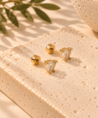 A pair of gold post earrings with pear-cut clear gemstones, displayed on a textured beige stone block with soft shadows and green leaves in the background.