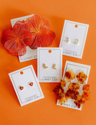 Five pairs of stud earrings displayed on white brand cards against a solid orange background. The earrings include large orange flower-shaped pieces, small gold heart studs, gold boot-shaped studs, and amber shell-and-floral stud sets.